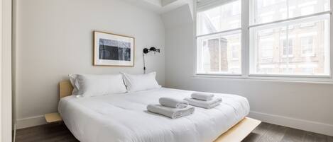 Una cama bien hecha con sábanas y toallas blancas, una lámpara de noche y un cuadro en la pared.