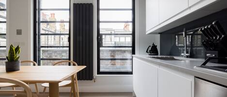 Een moderne keuken met witte kastjes, een houten eettafel en twee ramen met zwarte gordijnen.
