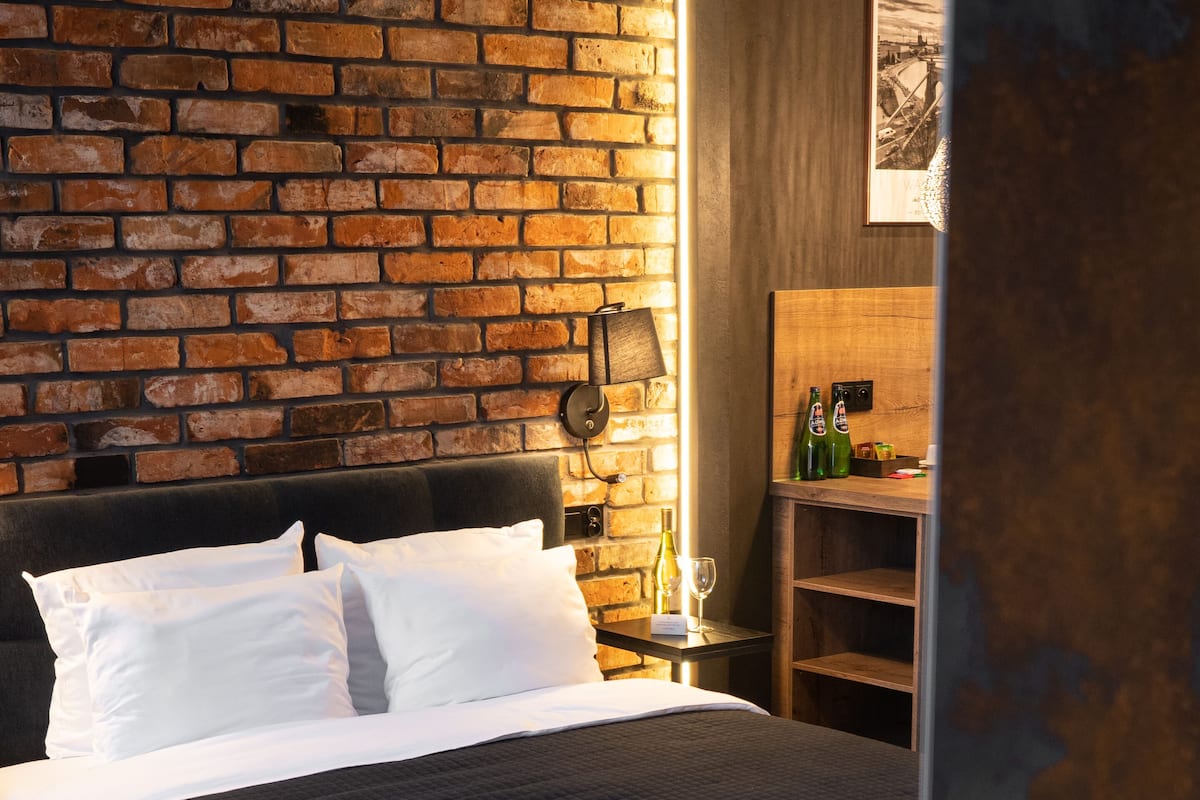 A hotel room with a brick wall, a bed with white linens, a nightstand with a lamp, and a small table with bottles and a glass.