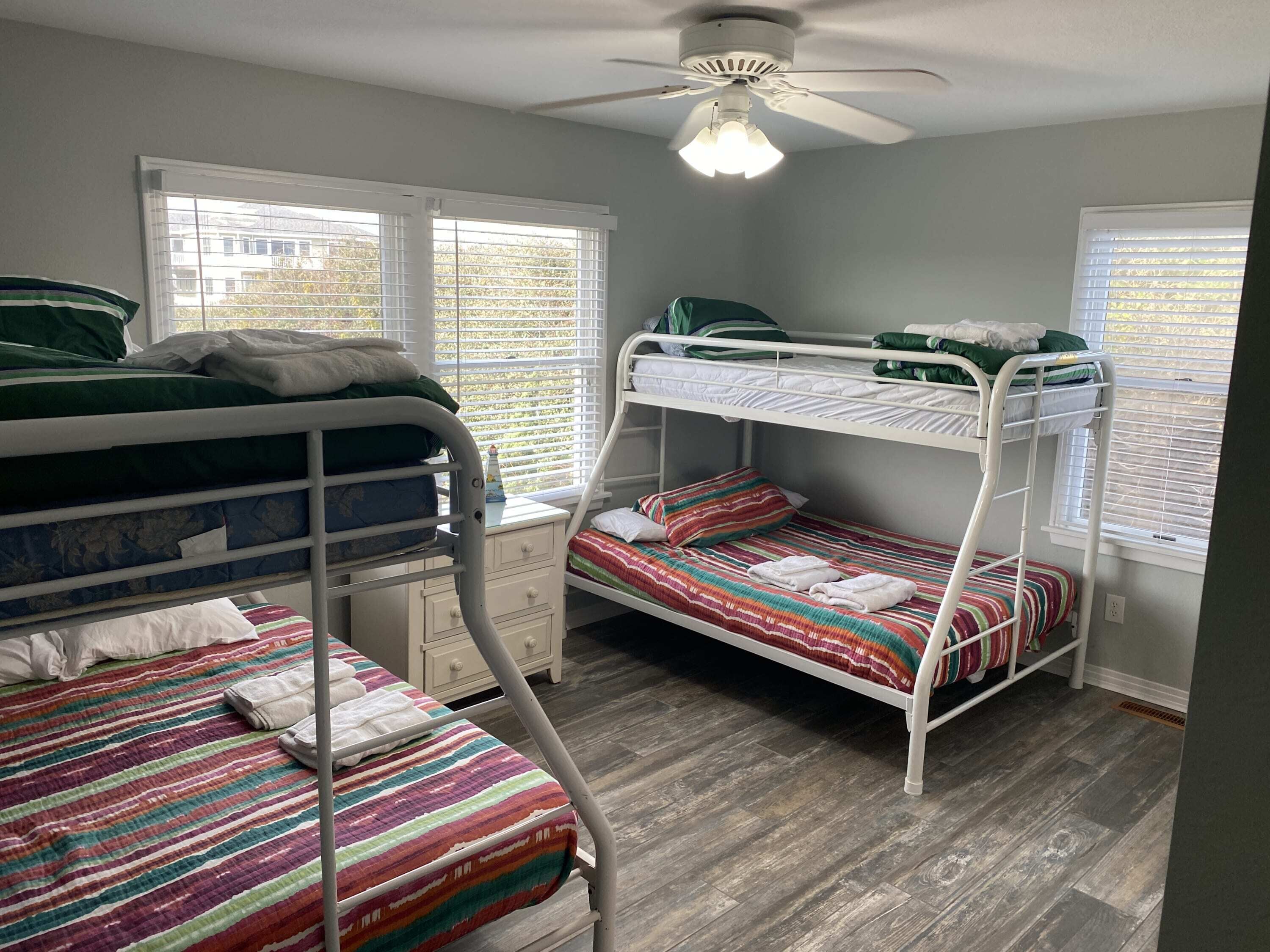 A bunk bed room with a ceiling fan, a window with blinds, and a wooden floor.