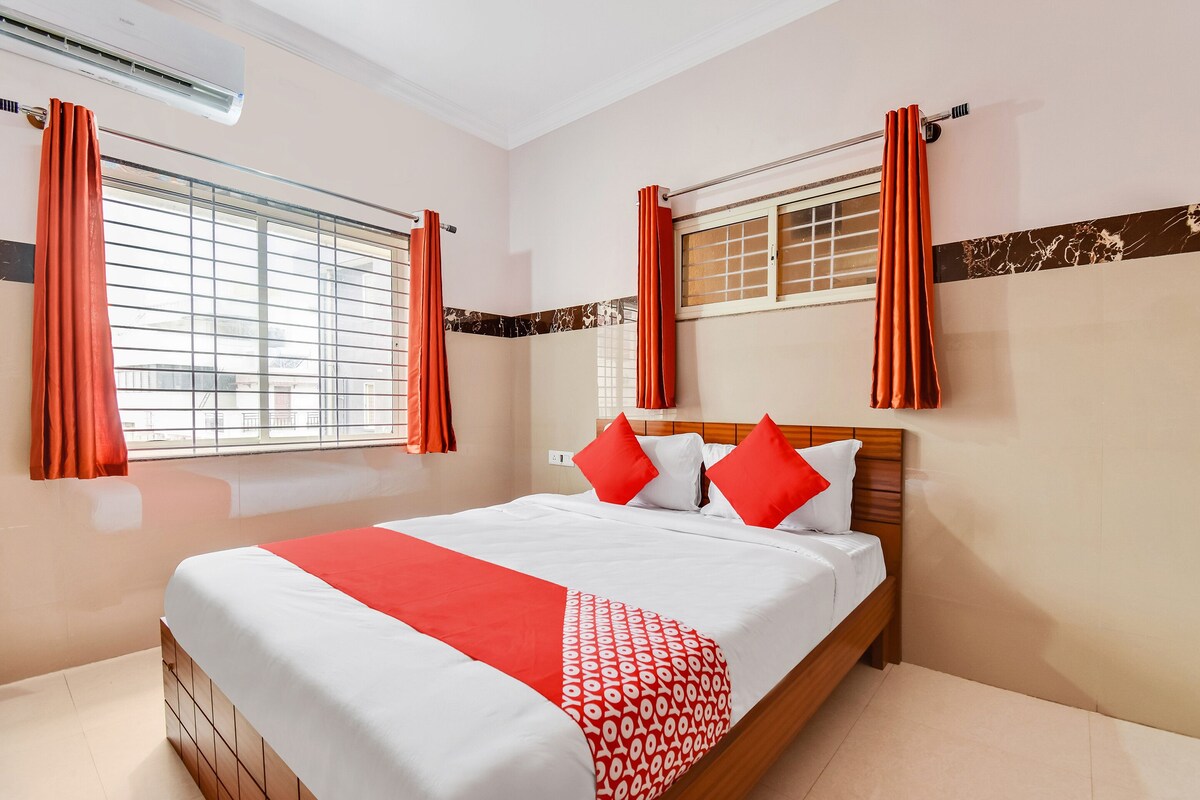 A hotel room with a large bed, two windows with curtains, a wooden headboard, and a red and white patterned bedspread.