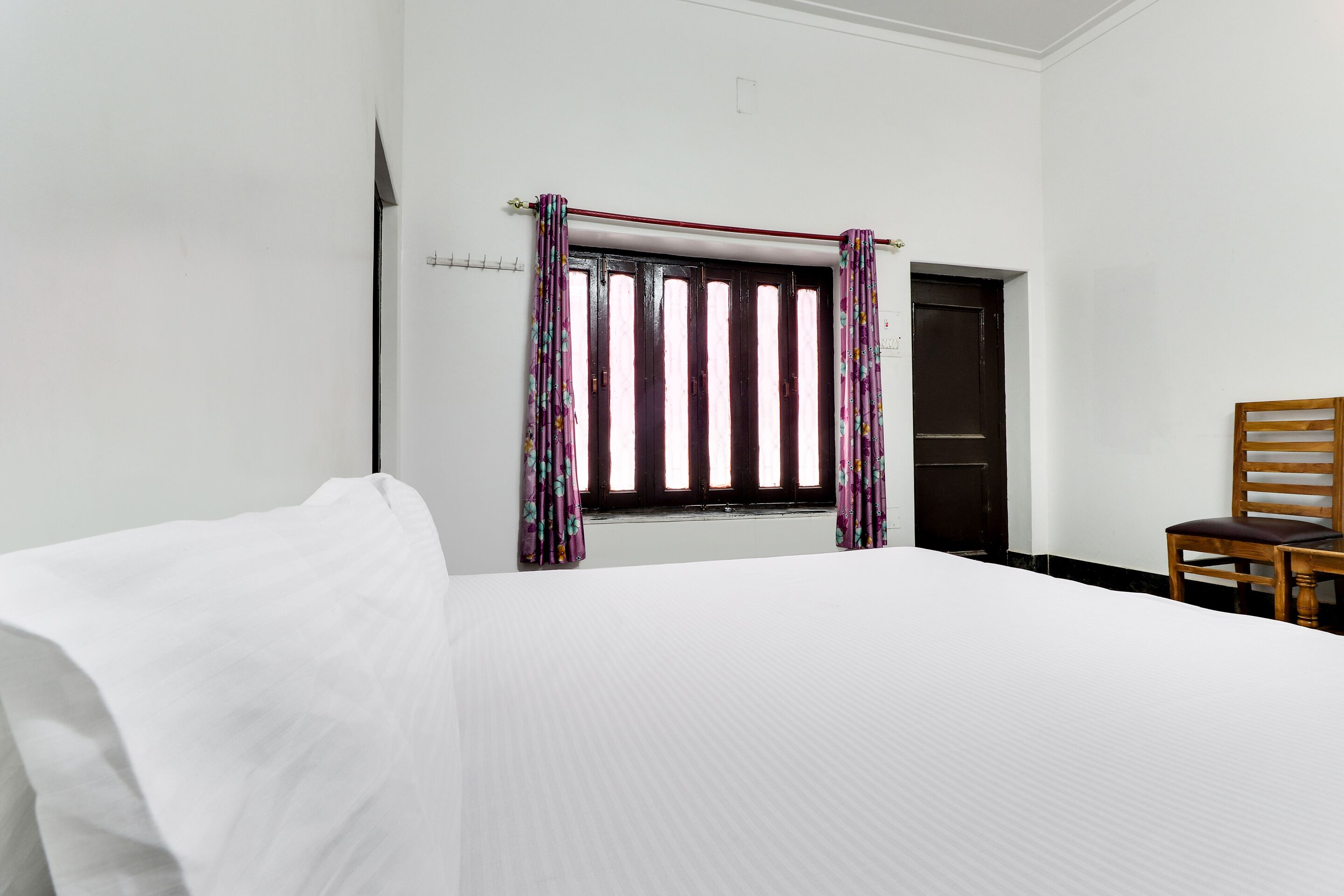 A hotel room with a bed, a window with curtains, a wooden chair, and a door.