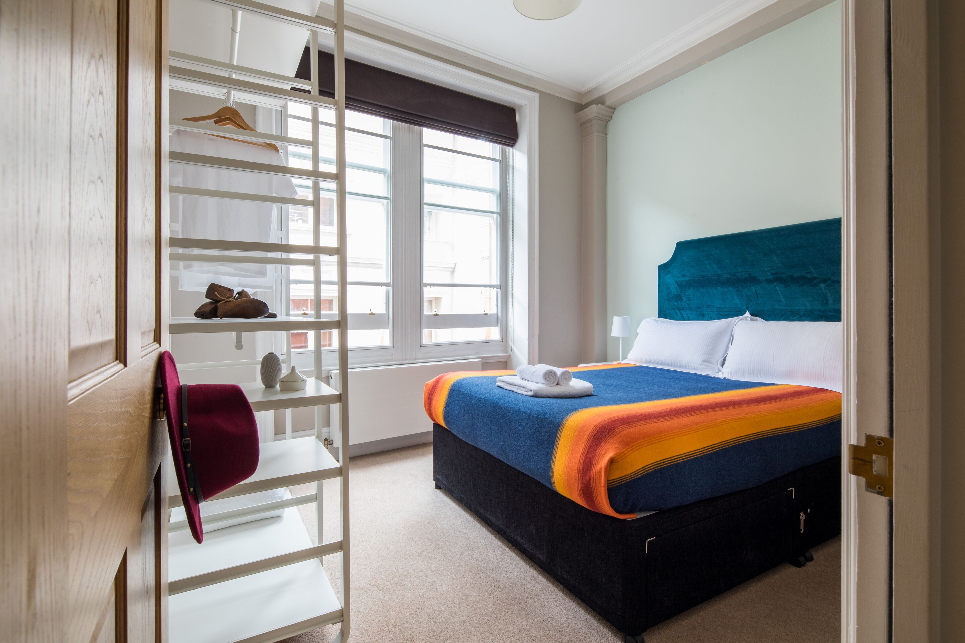 A bedroom with a bed, a window with blinds, a wardrobe, and a shoe rack.