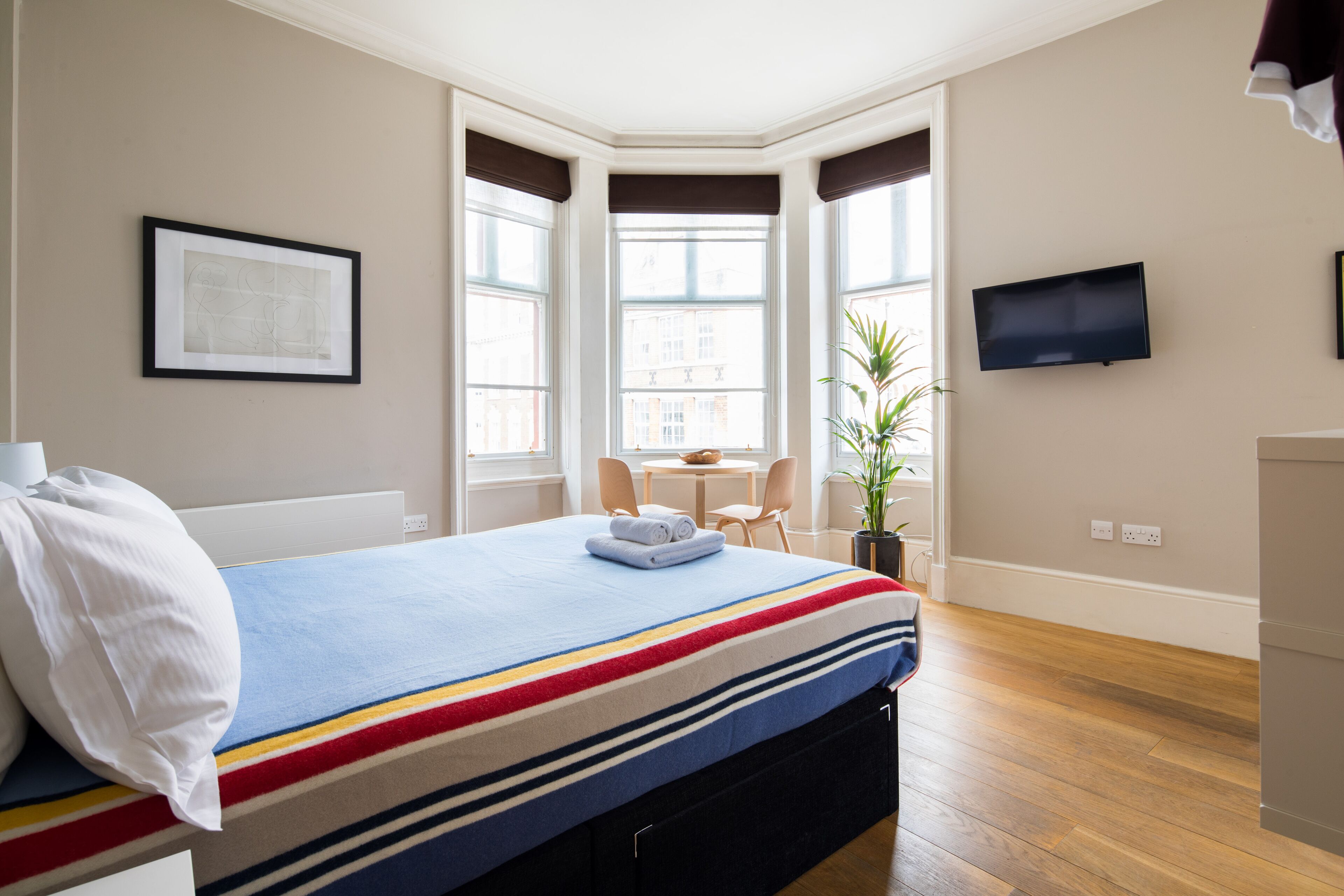 Une chambre d’hôtel avec un lit, une télévision, une fenêtre donnant sur des immeubles et une petite table avec des chaises.