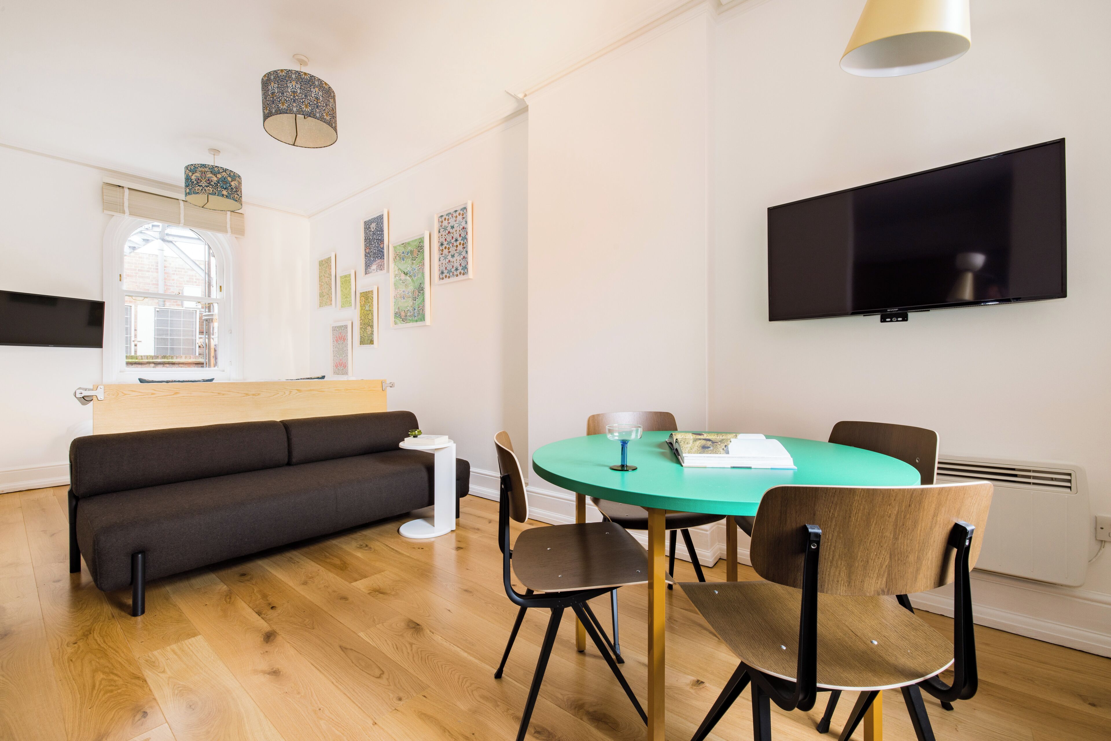 A modern living room with a sofa, a dining table with chairs, a flat-screen TV mounted on the wall, and artwork on the walls.