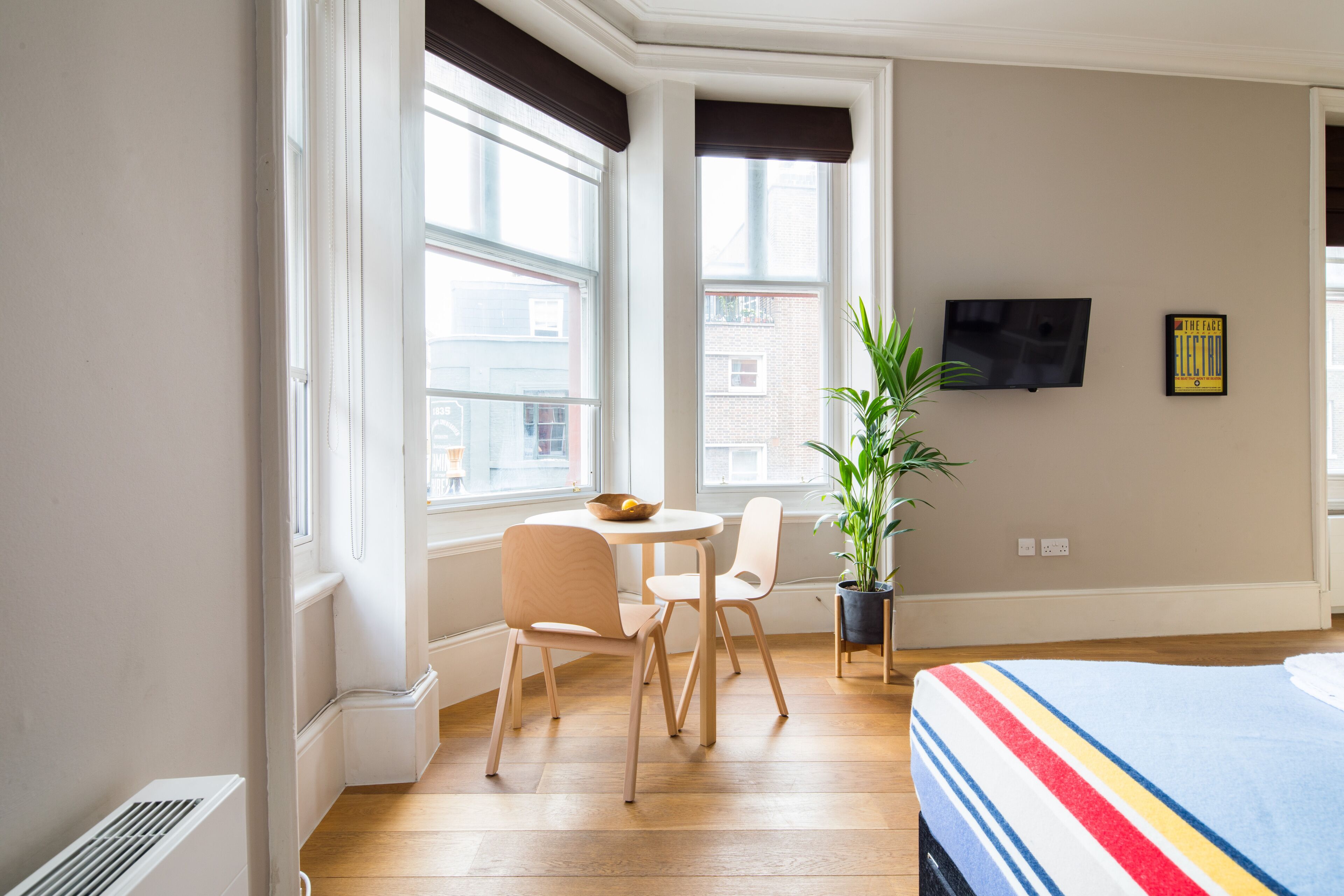 Une chambre d’hôtel moderne dotée d’une grande fenêtre, d’une table ronde avec des chaises, d’une plante en pot, d’un téléviseur à écran plat et d’un lit recouvert d’une couverture à rayures.