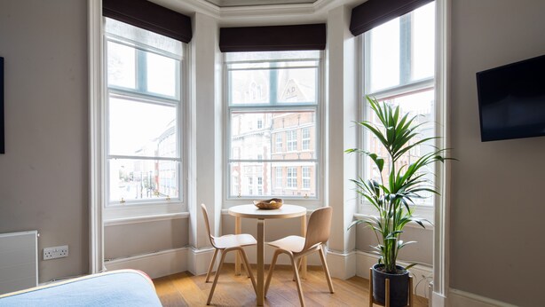 A room with a bed, a round table with two chairs, a potted plant, and large windows with a view of buildings. - Hausd - Covent Garden (London)