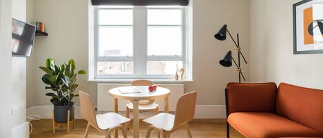 A modern living room with a round table, two chairs, a sofa, a potted plant, and a large window with a view of the city.