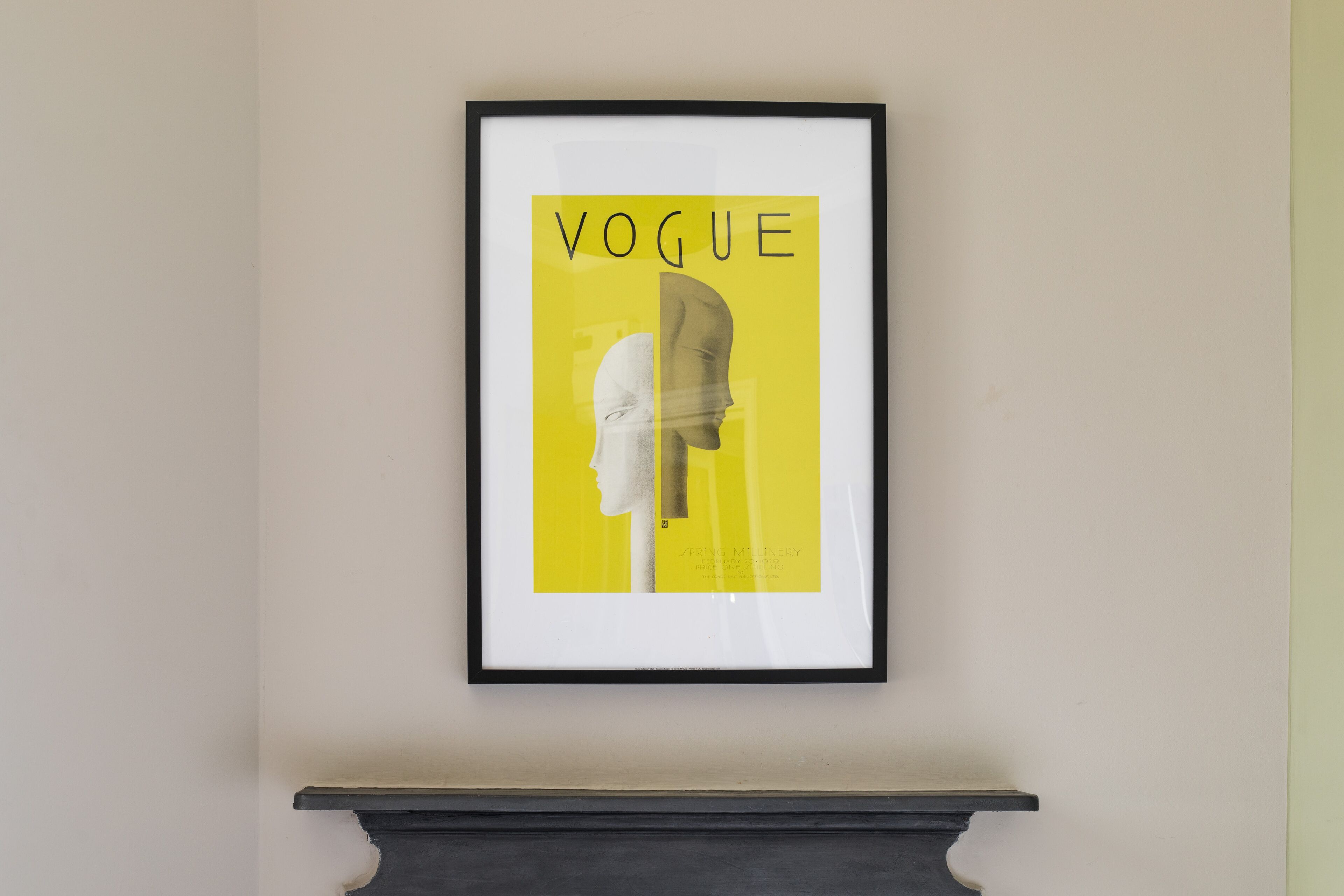 A framed artwork with the word "VOGUE" on a yellow background, mounted on a wall above a dark mantelpiece.