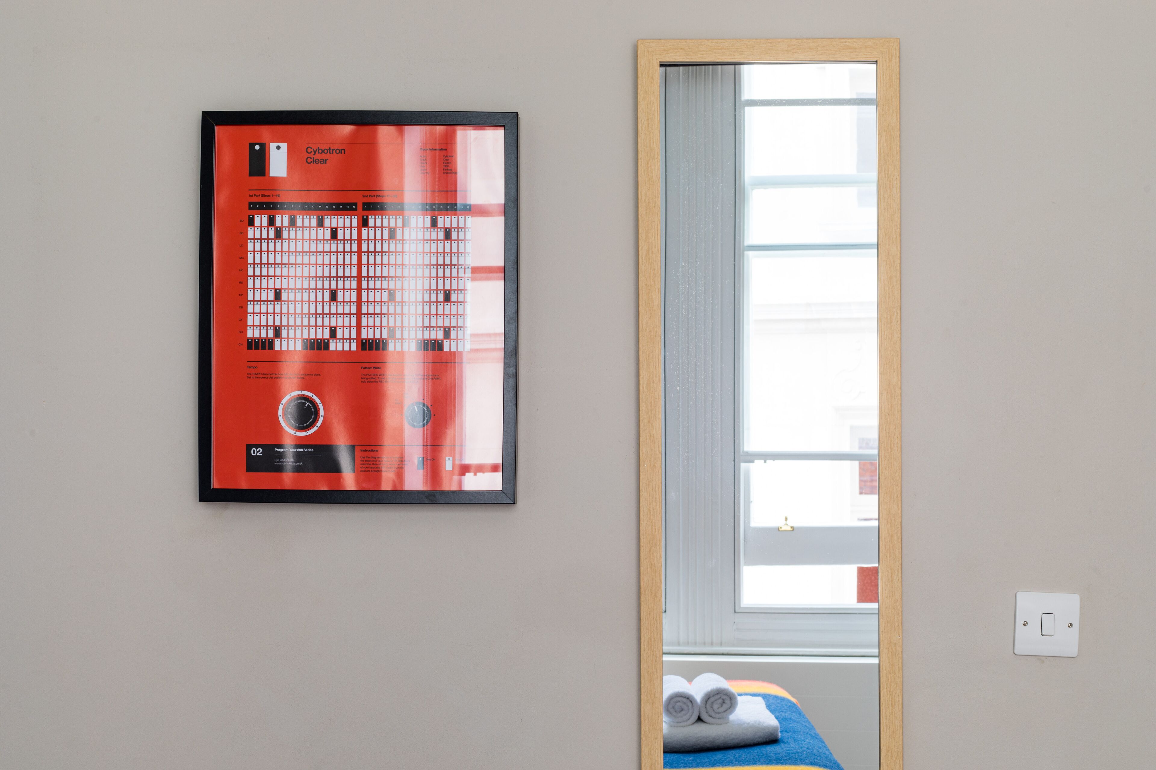 A framed chart on the wall, a window with blinds, and a bed with rolled towels.