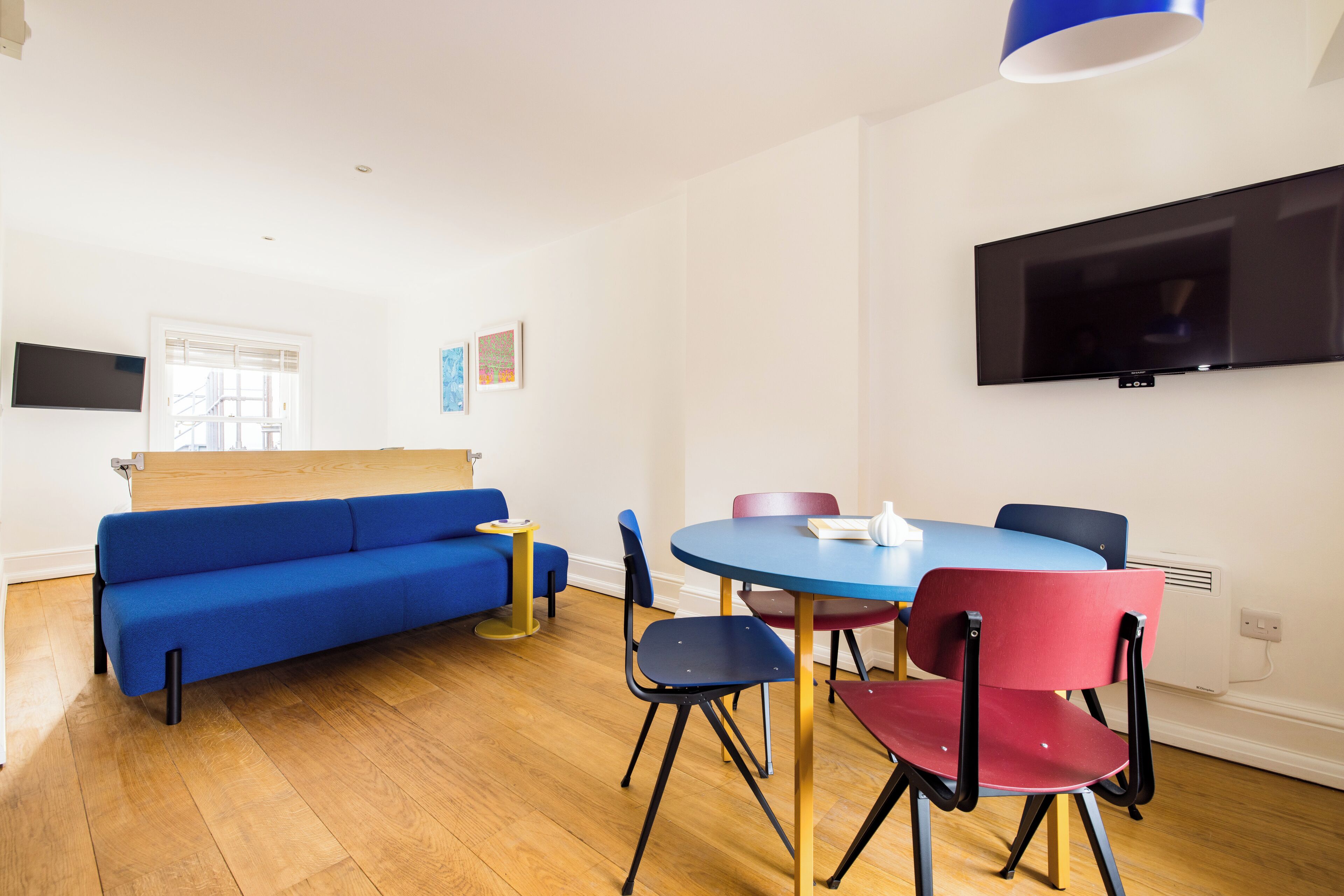 Un salon moderne avec un canapé bleu, une table ronde entourée de chaises, un téléviseur à écran plat fixé au mur et une fenêtre équipée de stores.