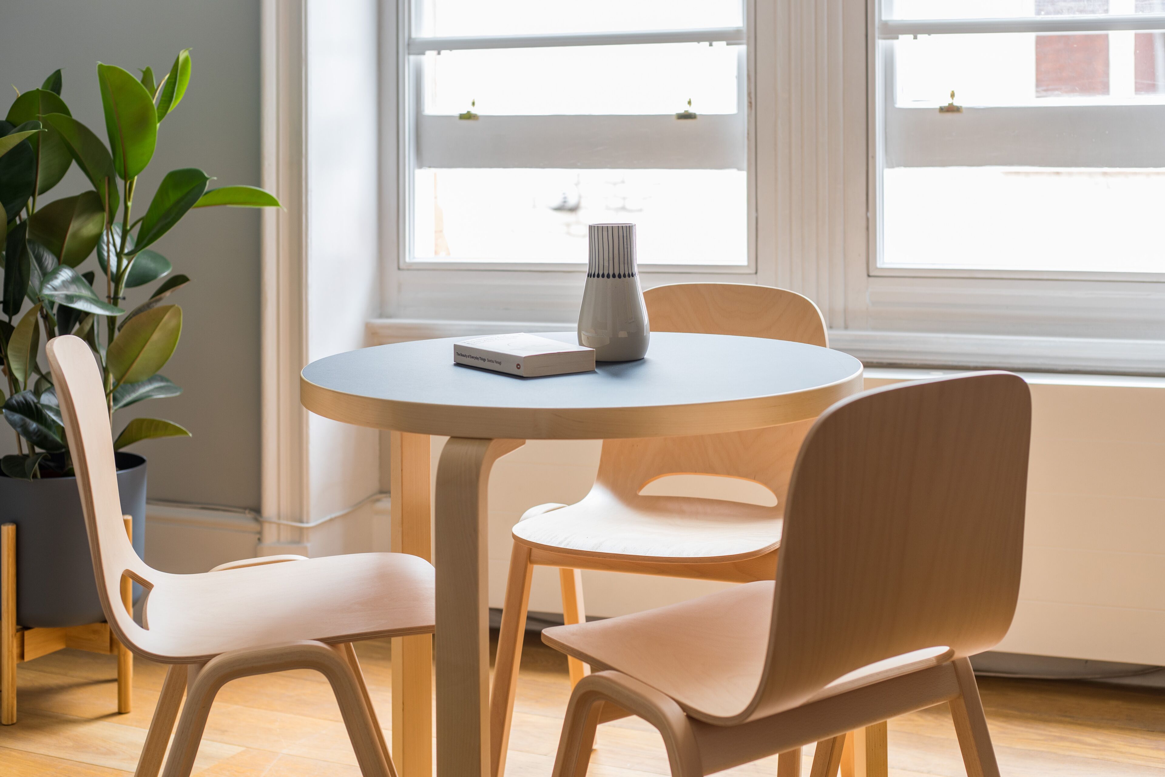 Ein runder Tisch mit Buch und Blumenvase, umgeben von zwei Stühlen, in einem Zimmer mit großem Fenster und Topfpflanze.