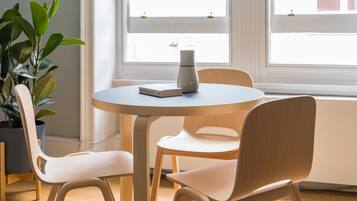 Ein runder Tisch mit Buch und Blumenvase, umgeben von zwei Stühlen, in einem Zimmer mit großem Fenster und Topfpflanze.