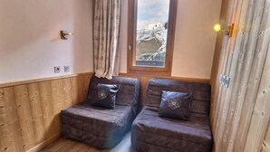 Une pièce chaleureuse avec deux canapés marron foncé, chacun agrémenté d’un coussin décoratif, des lambris en bois aux murs, une fenêtre donnant sur des montagnes enneigées et un luminaire fixé au mur.