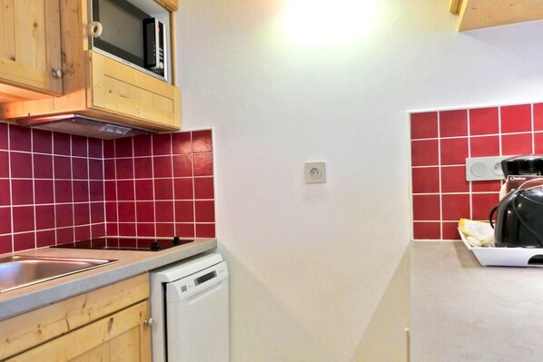 A compact kitchen with wooden cabinets, a microwave, a dishwasher, and red-tiled walls.