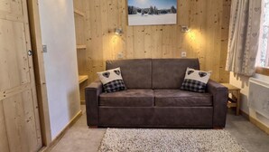 A cozy room with a brown leather sofa, two heart-patterned pillows, a small wooden table, and a painting of a snowy landscape on the wall. - 10m from ski slopes, Ground floor, view limited, 27m², Méribel-Mottaret (Méribel - Les Allues)
