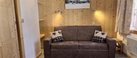 A cozy room with a brown leather sofa, two heart-patterned pillows, a small wooden table, and a painting of a snowy landscape on the wall.