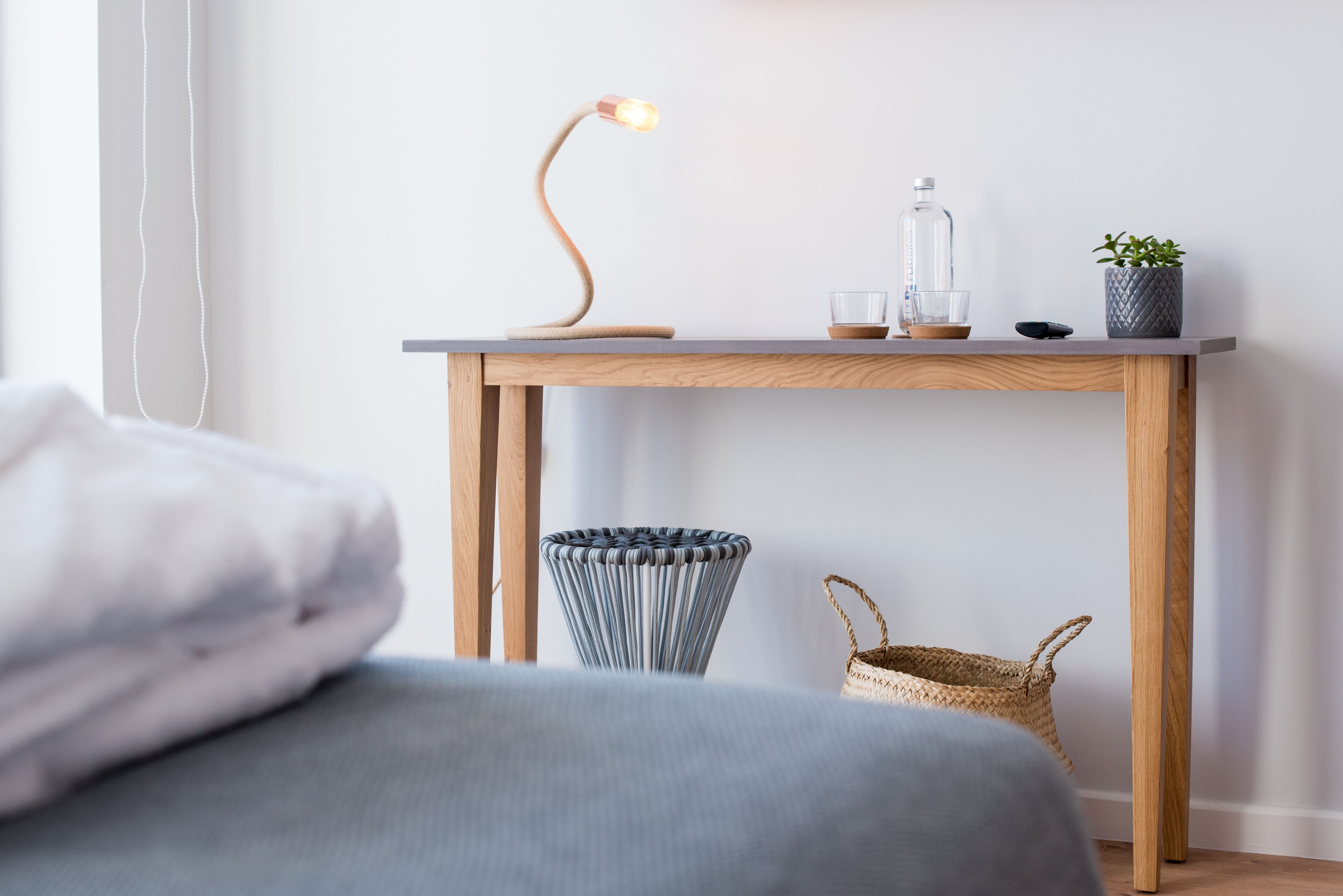 Una camera da letto ordinata con un comodino in legno, un tavolino, una lampada, una bottiglia di vetro, una pianta in vaso e un cesto di vimini.