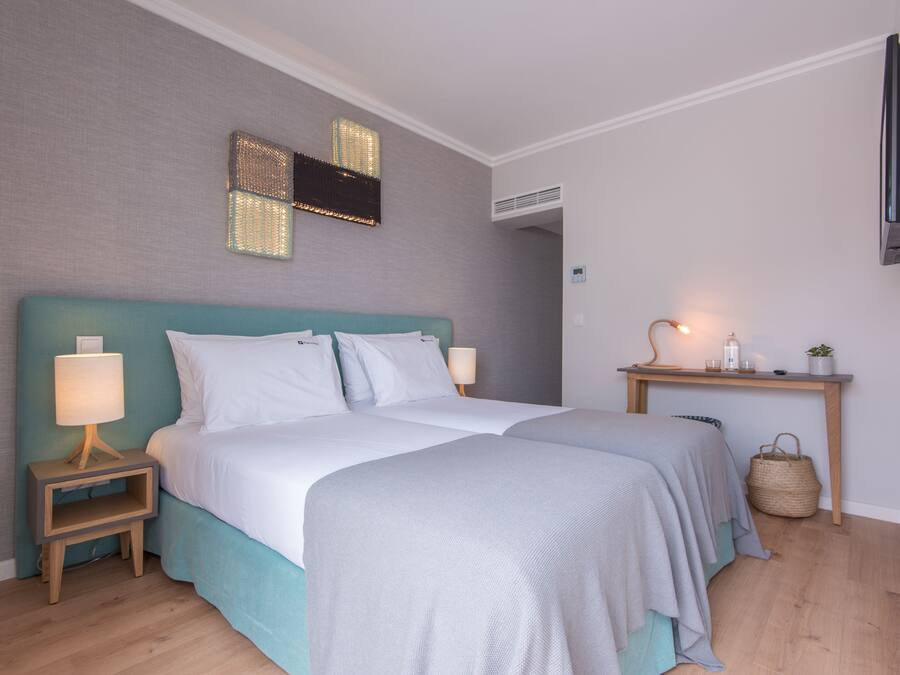 A double bed with a teal headboard, two bedside tables with lamps, a flat-screen TV mounted on the wall, and a small bedside table with a basket.
