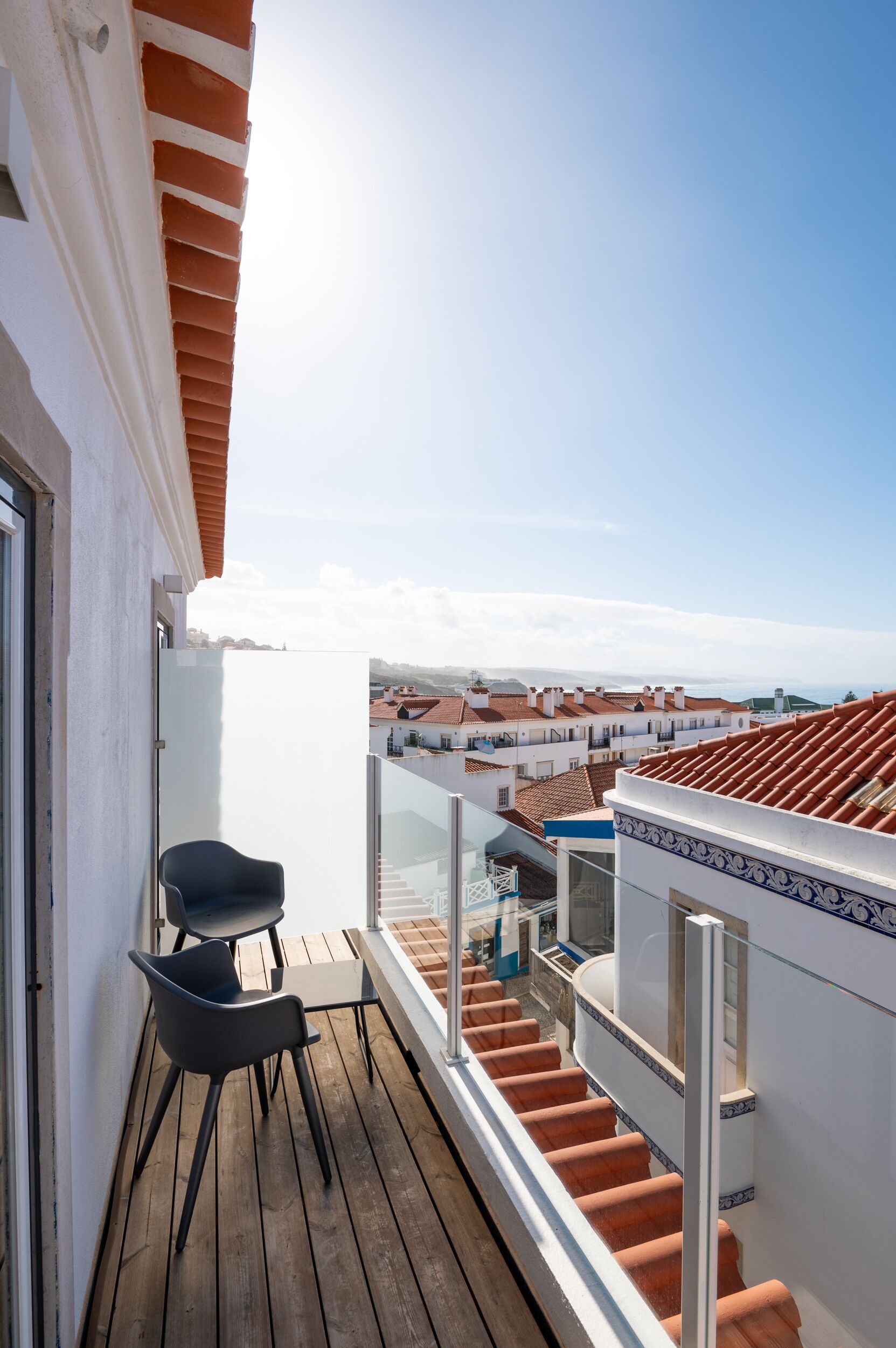 A balcony with a wooden floor and two black chairs, overlooking a cityscape with terracotta roofs.
