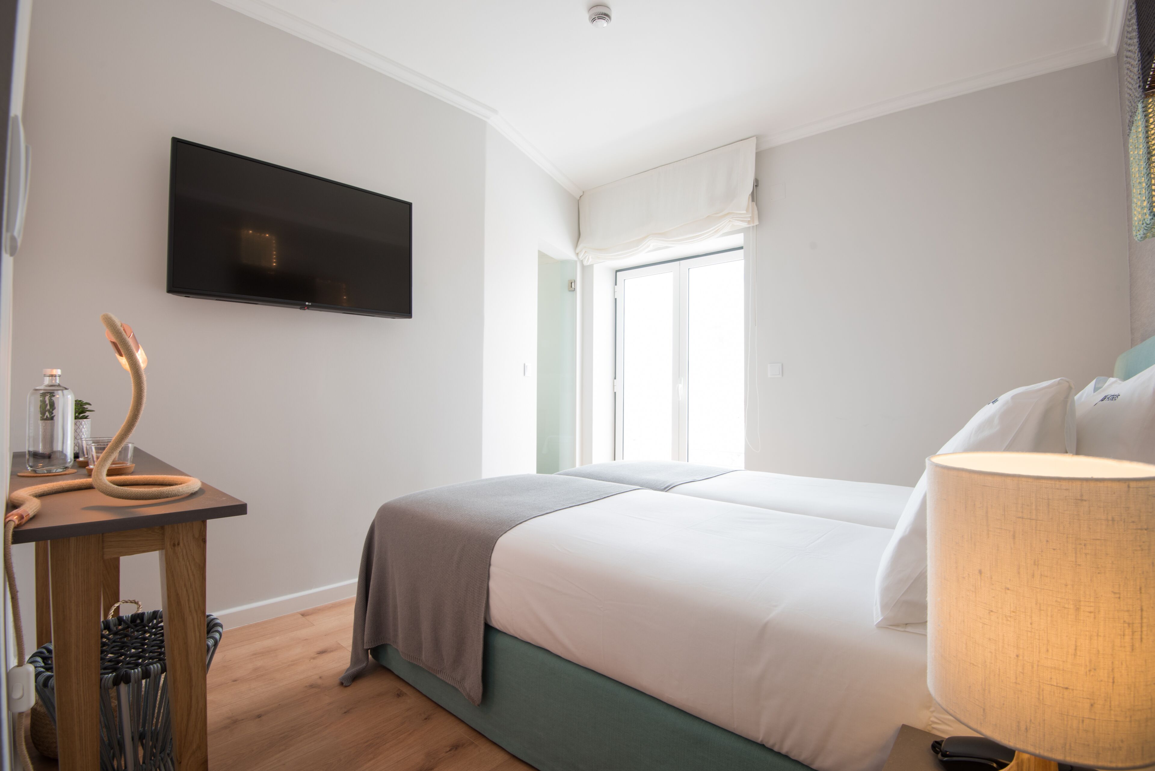 A hotel room with a bed, a TV mounted on the wall, a window with curtains, a bedside table with a lamp, and a chair.