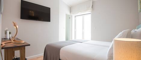 A hotel room with a bed, a TV mounted on the wall, a window with curtains, a bedside table with a lamp, and a chair.