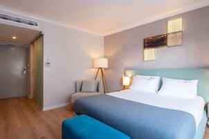 Quarto de hotel com cama, mesa de cabeceira, luminária, cadeira e iluminação fixa na parede.