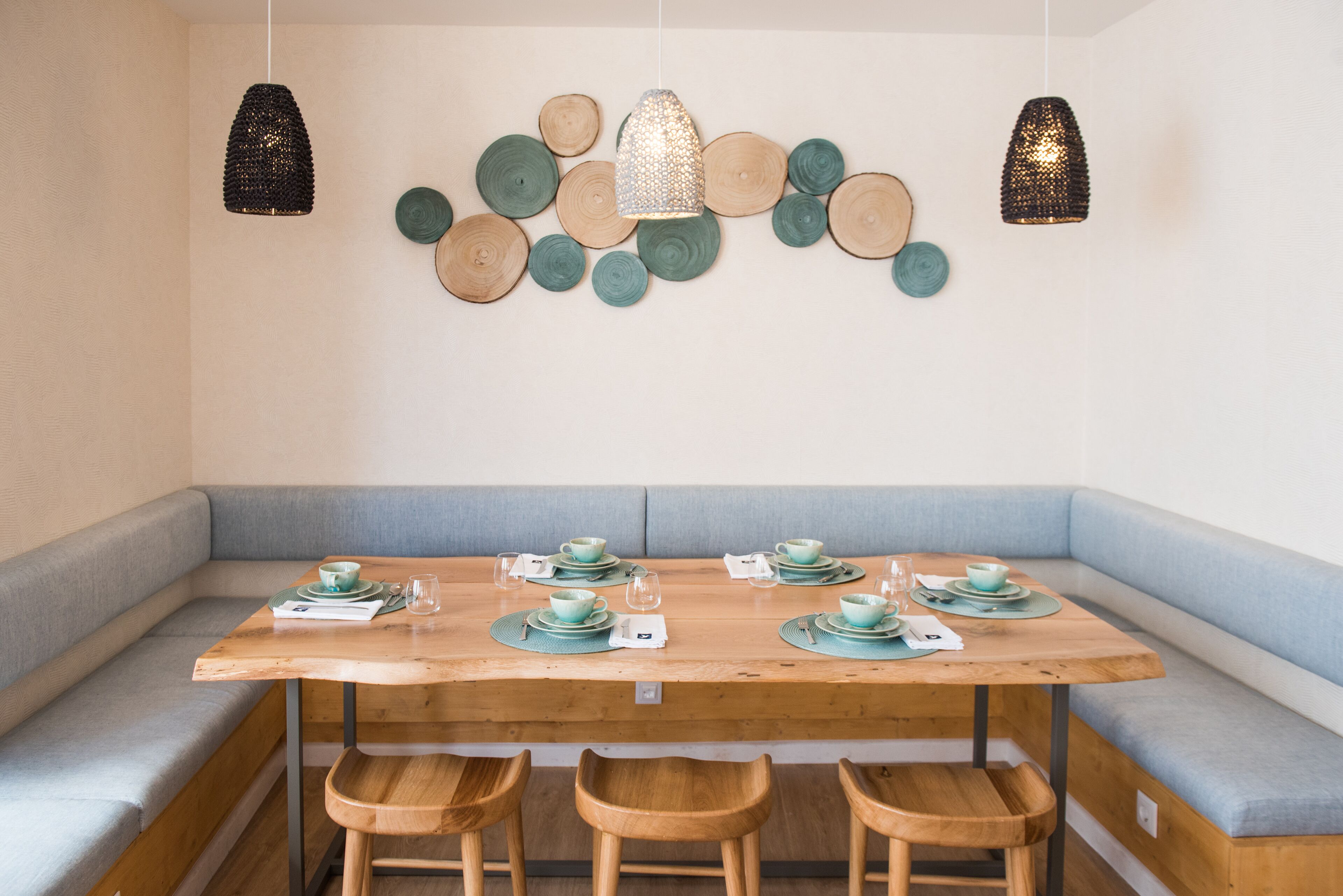 Un comedor con una mesa de madera dispuesta para seis personas, bancos acolchados azul claro y lámparas colgantes.