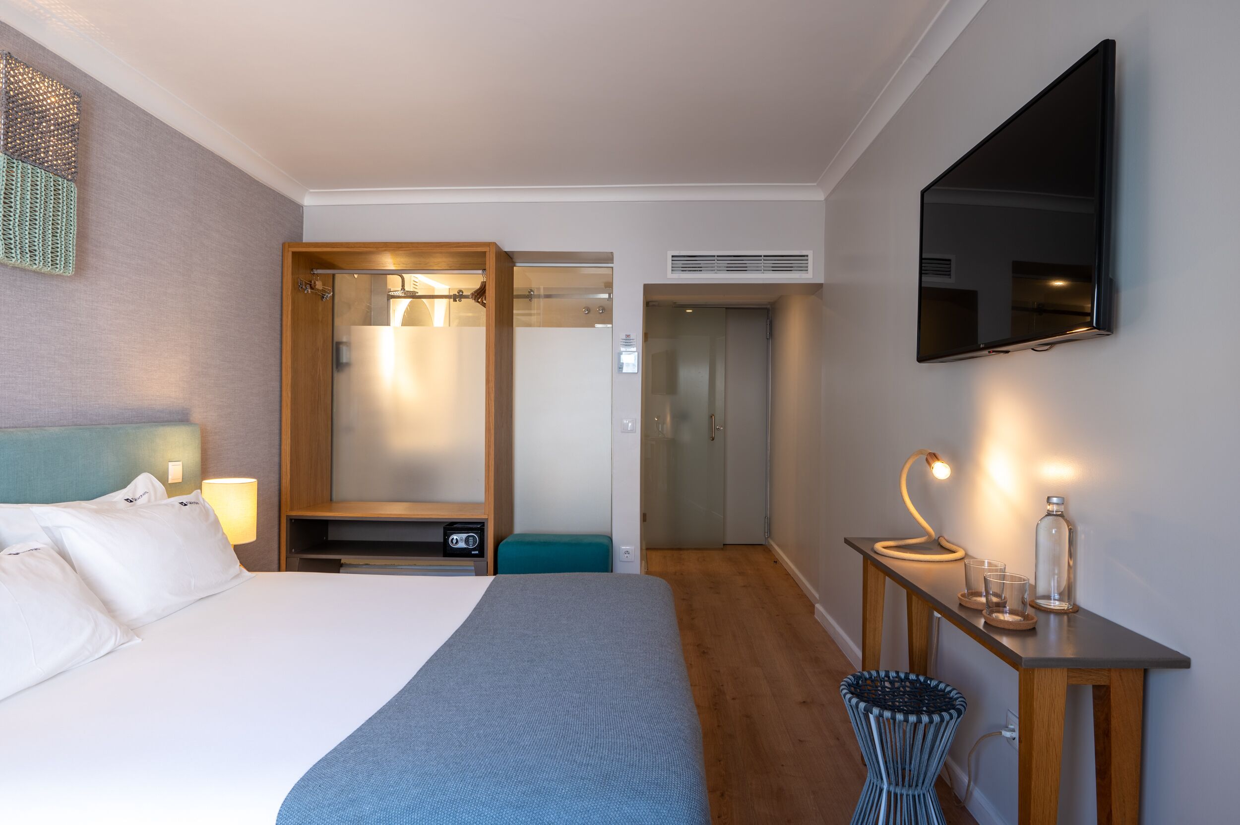 A hotel room with a bed, bedside lamp, TV, and a small table with glasses and a bottle.