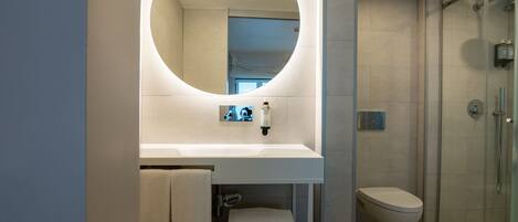 A modern bathroom with a round mirror, a white sink, a glass shower enclosure, and towels with the FLH Hotels logo.