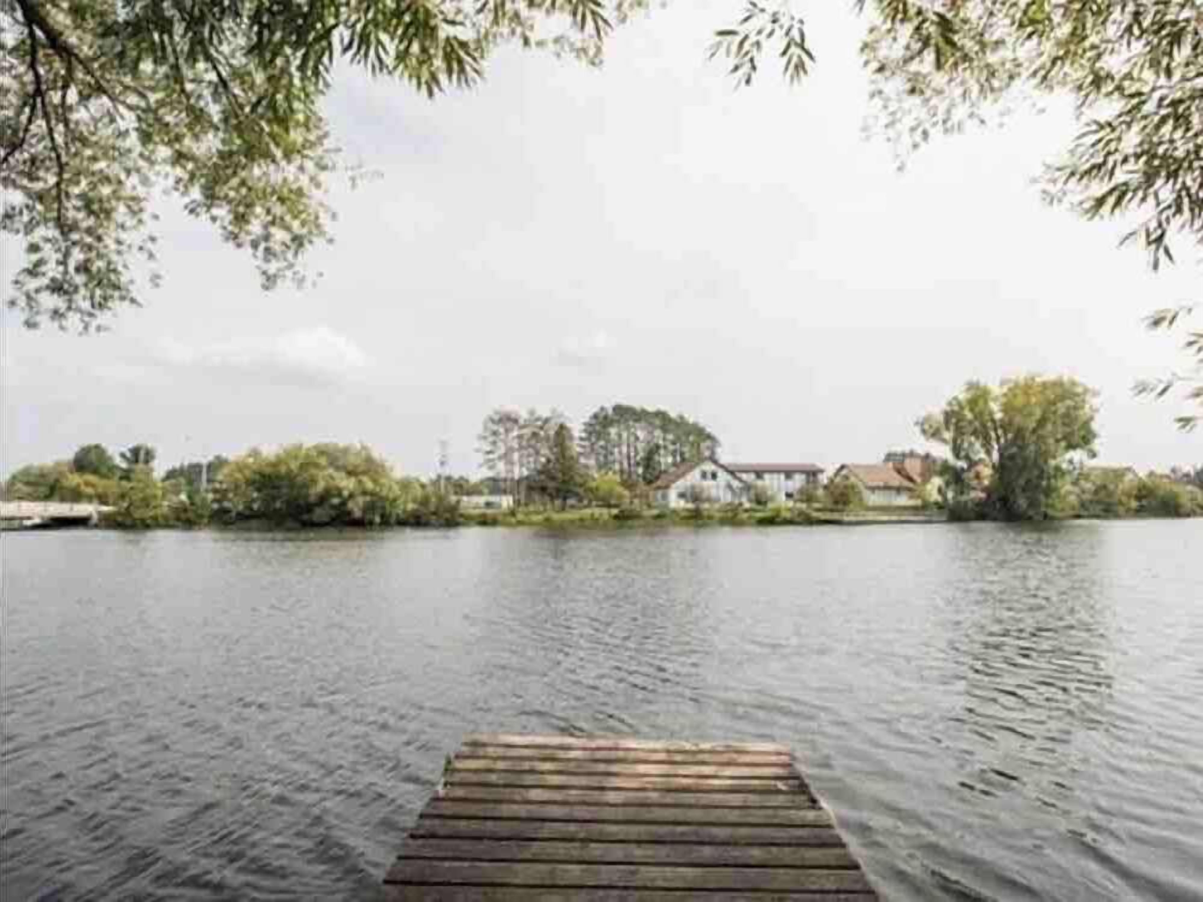 Eine Holzstege reicht in einen stillen See, im Hintergrund sind Gebäude und Bäume zu sehen.