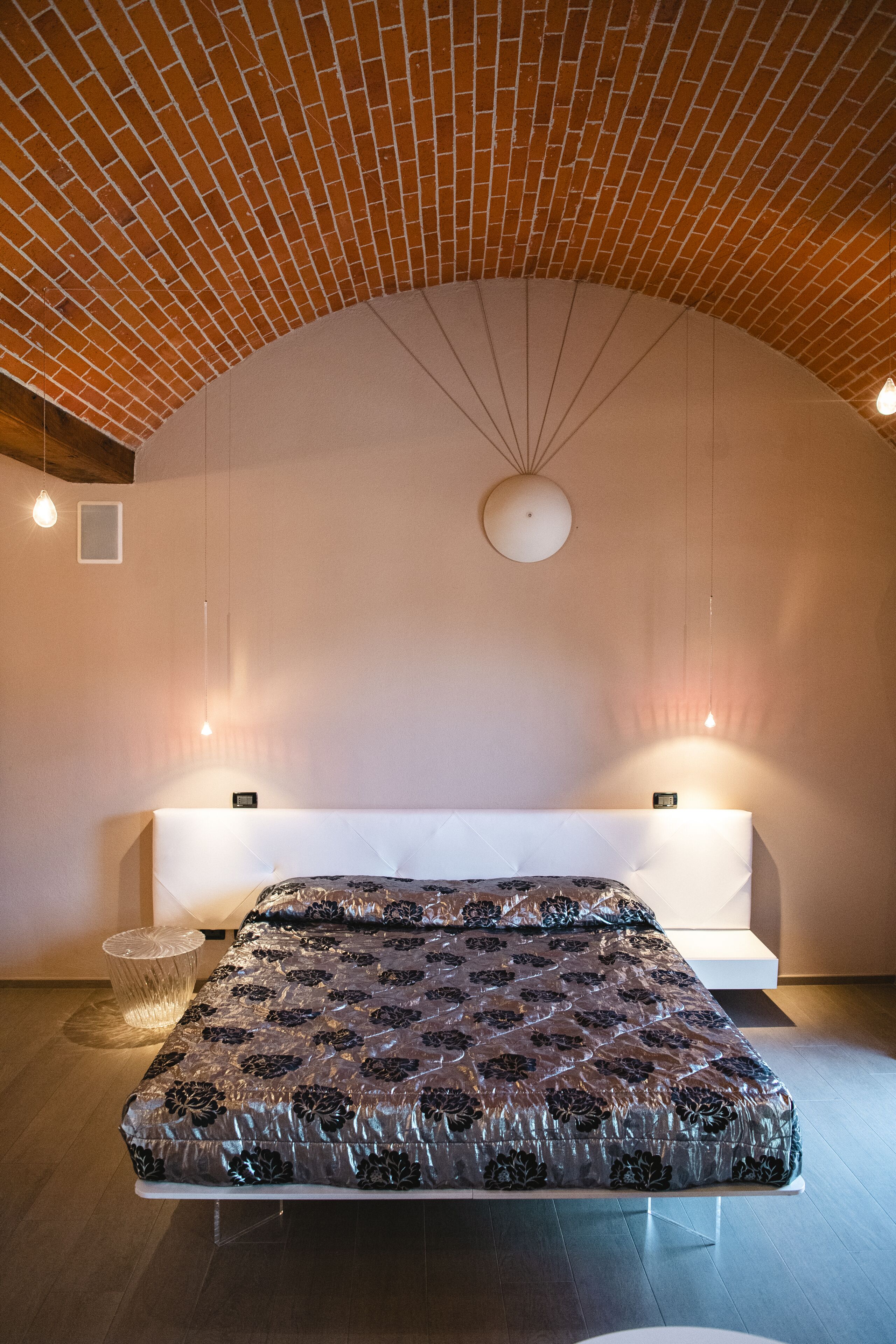 A modern bedroom with a brick ceiling, a large bed with a patterned bedspread, a white bench, and pendant lighting.