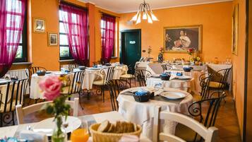 Un espace repas avec plusieurs tables préparées pour le petit-déjeuner, où l’on trouve du pain, des fruits et des boissons.