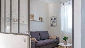 Un salon moderne avec un canapé gris, une petite table ronde ornée d’un pot de plante, et un mur blanc décoré d’un motif quadrillé et de photos encadrées.