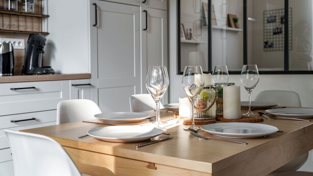 Un espace repas moderne avec une table en bois dressée pour quatre personnes, des chaises blanches et une cuisine en arrière-plan.
