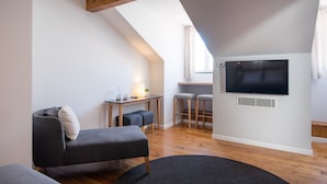 A modern loft bedroom with a sloped ceiling, a large TV mounted on the wall, a desk with two stools, and a cozy seating area with a sofa and ottoman. - Urbano FLH Hotels Lisboa (Lisbon)