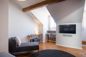 Un dormitorio tipo loft moderno con techo inclinado, un televisor grande montado en la pared, un escritorio con dos taburetes y una zona de estar acogedora con sofá y reposapiés.