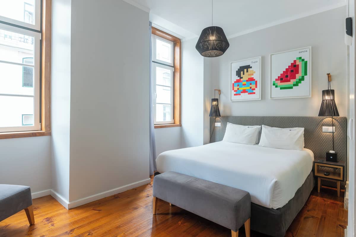 Une chambre à coucher moderne avec un grand lit, un banc, une table de chevet avec une lampe et des œuvres d’art accrochées au mur.