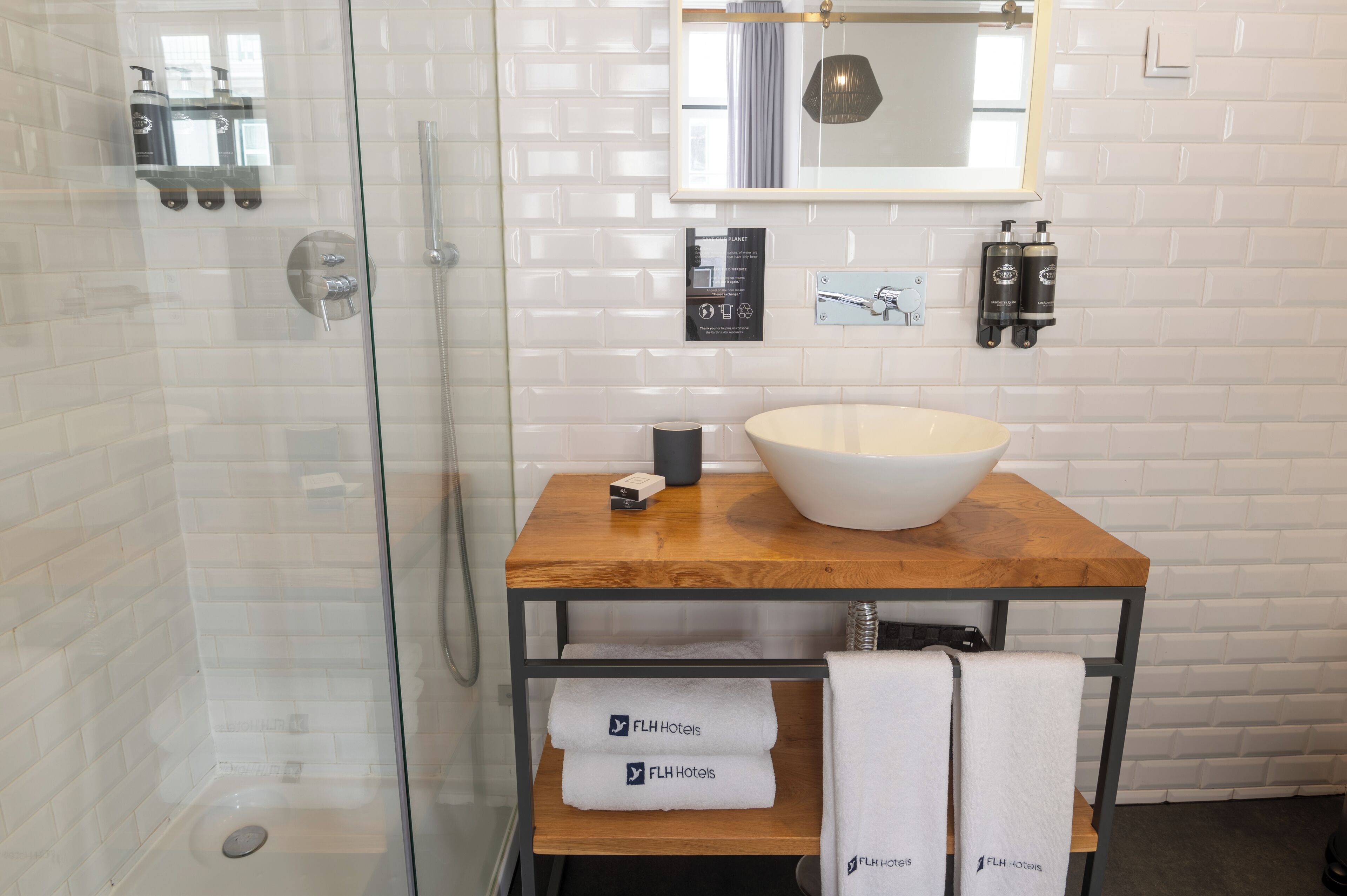 A modern bathroom with a glass shower enclosure, a wooden vanity with a large white basin, and a mirror above the sink. There are two bottles of shampoo on the wall and a towel rack with towels.