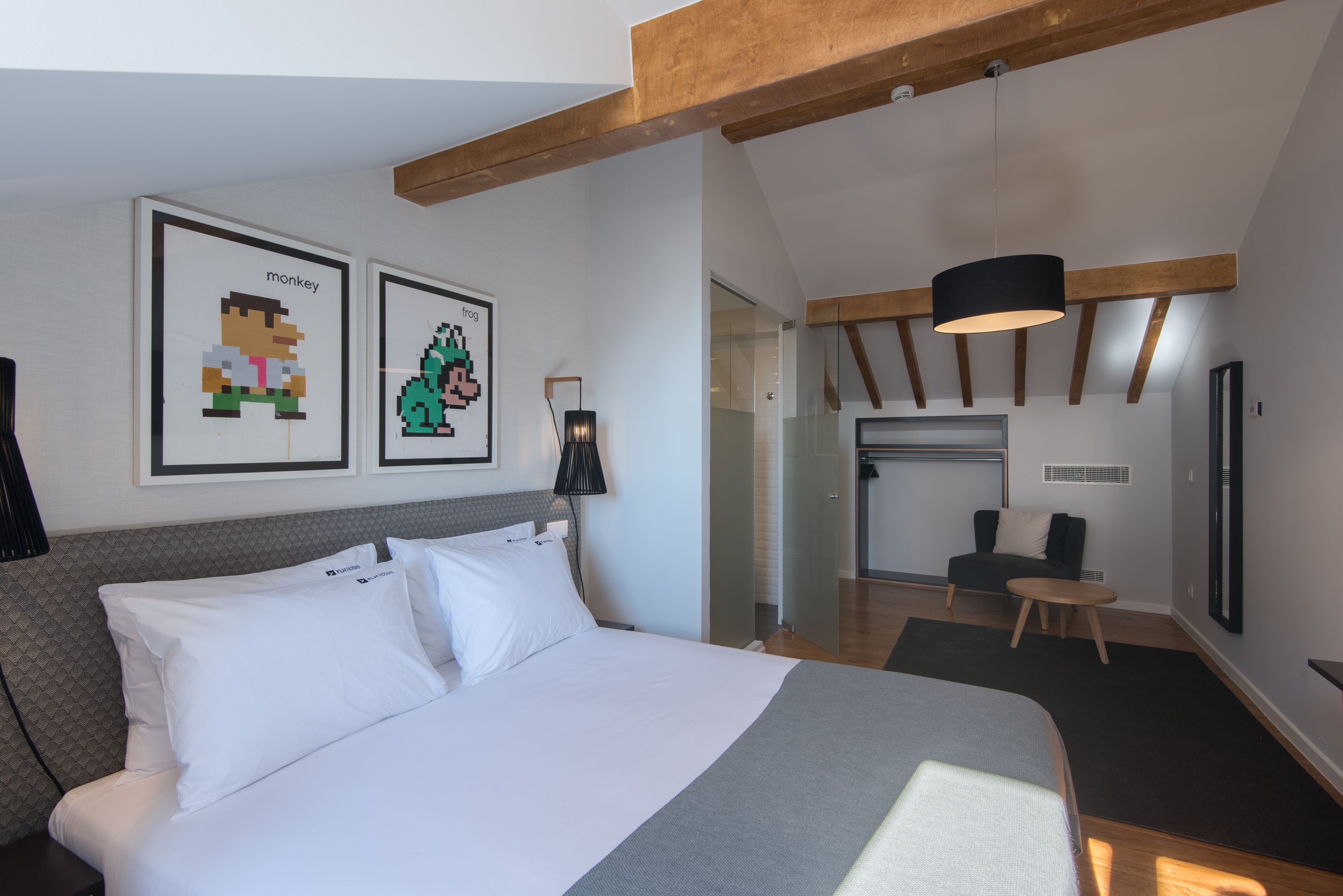 A modern hotel room with a large bed, a desk, and a chair. There are two framed pixel art pictures on the wall.