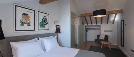 A modern hotel room with a large bed, a desk, and a chair. There are two framed pixel art pictures on the wall.
