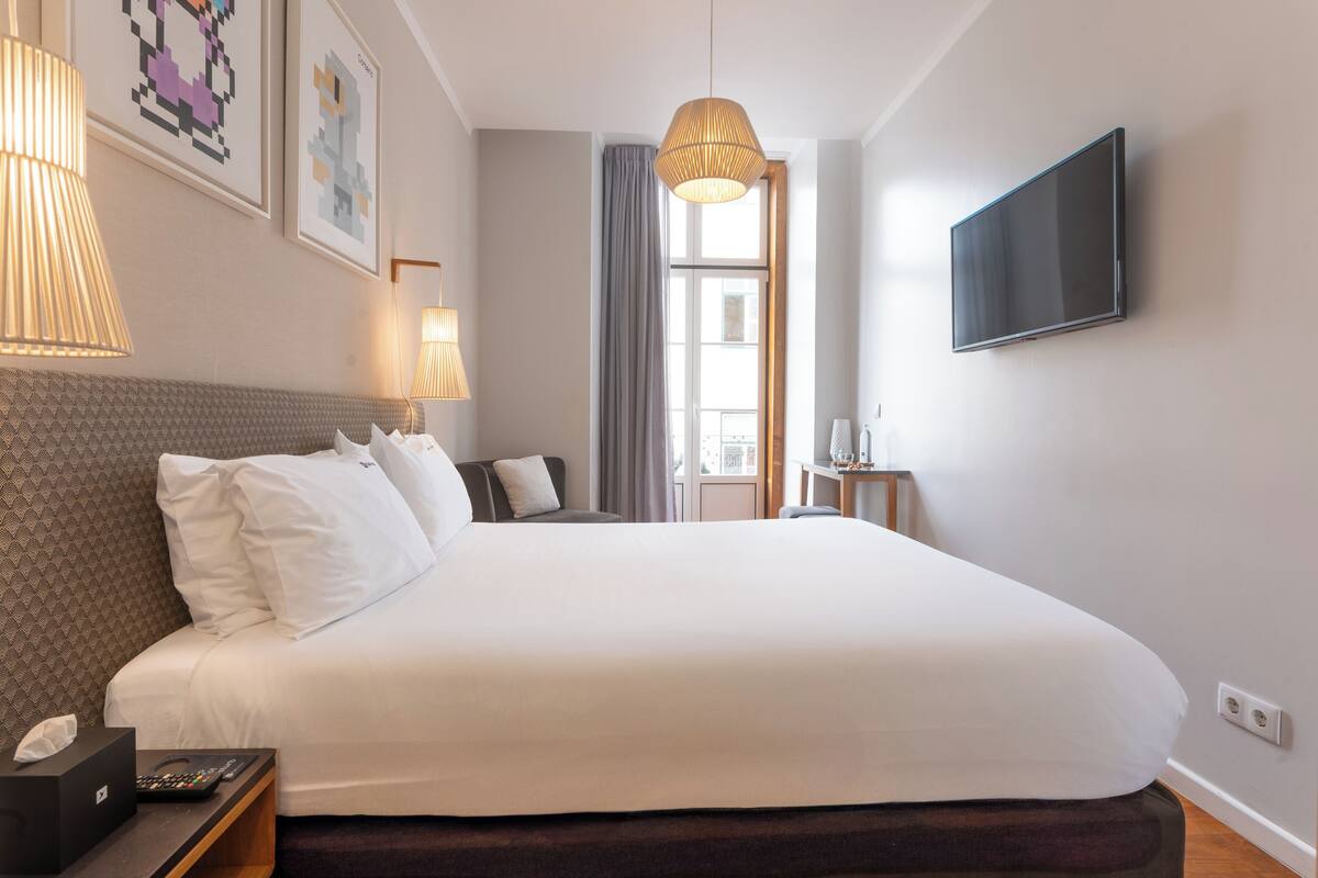 Un lit bien fait, avec du linge de lit blanc, une tête de lit et une table de chevet sur laquelle se trouvent une lampe et une télécommande.