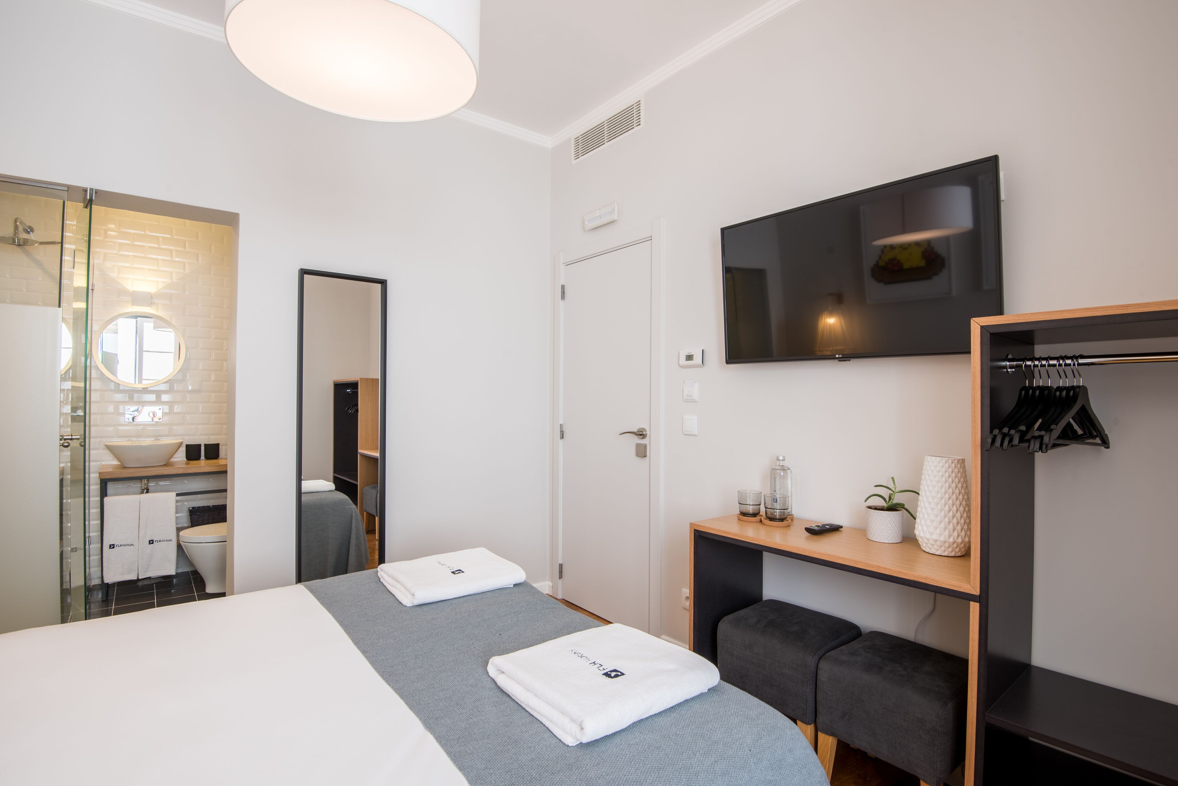 Ein modernes Hotelzimmer mit Bett, zwei Nachttischen, Fernseher, Spiegel, Kleiderschrank und einem Badezimmer mit Waschbecken und Toilette.