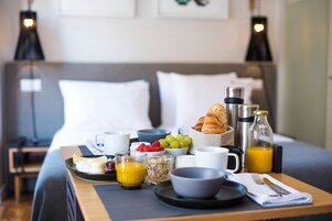 Una bandeja de desayuno ordenada sobre la cama, con un almohadón, una mesita de noche con una lámpara y un vaso de zumo de naranja.