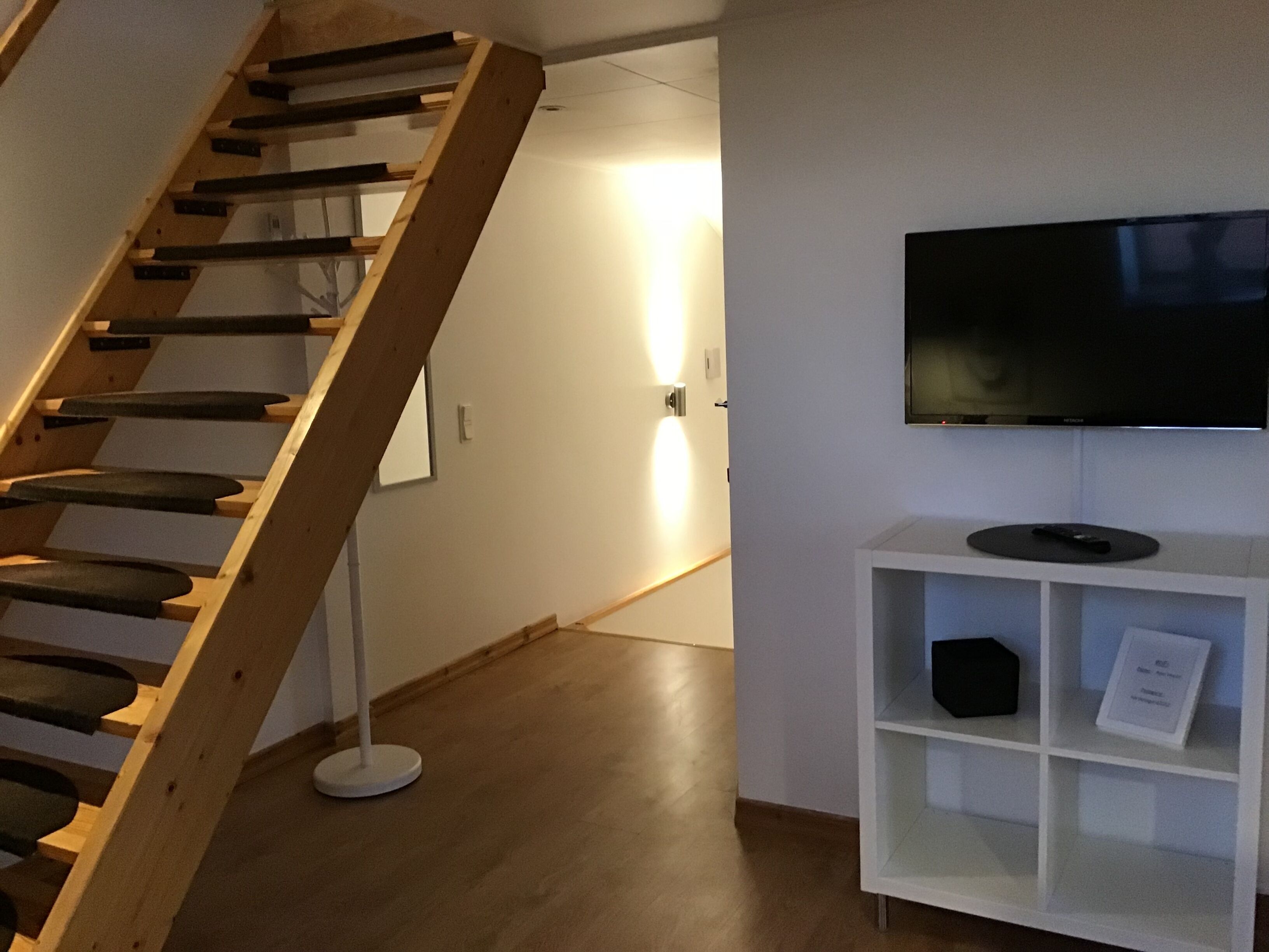 Una escalera con barandilla de madera que lleva a una pequeña plataforma. Se ve un estante blanco con un televisor y un control remoto.
