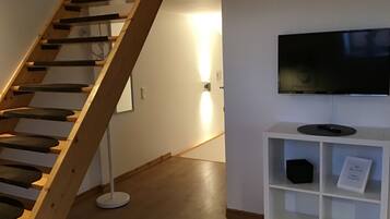 Una escalera con barandilla de madera que lleva a una pequeña plataforma. Se ve un estante blanco con un televisor y un control remoto.