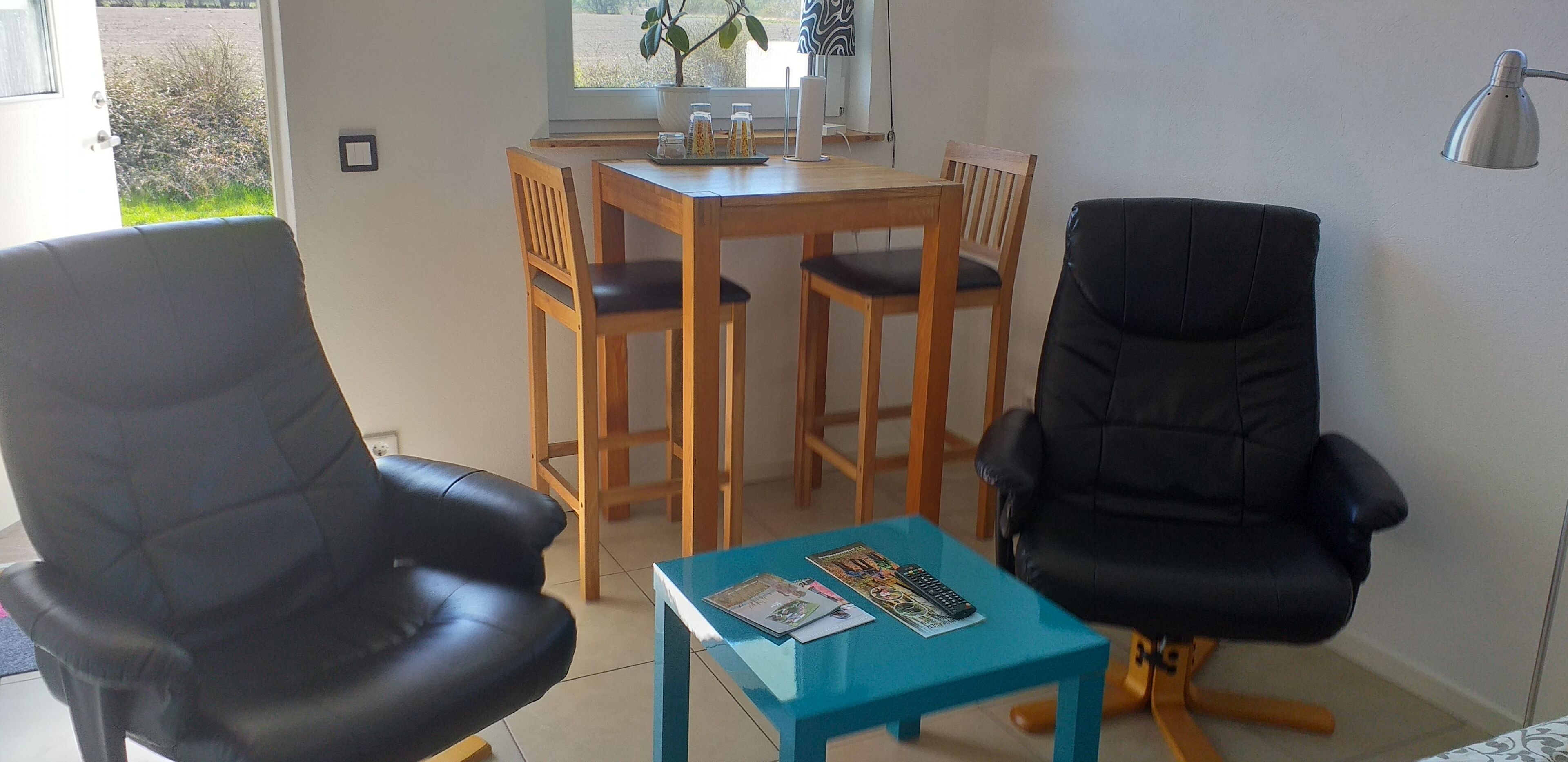 A living room with two black leather chairs, a wooden dining table with four chairs, a small blue coffee table, and a potted plant by the window.