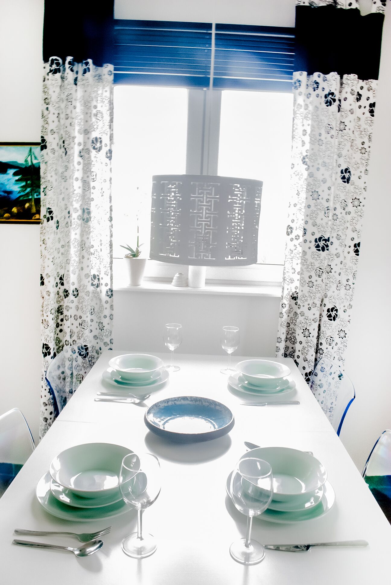 Une table à manger dressée avec des assiettes, des verres et des couverts, encadrée de rideaux à motifs et d’une fenêtre avec des stores.