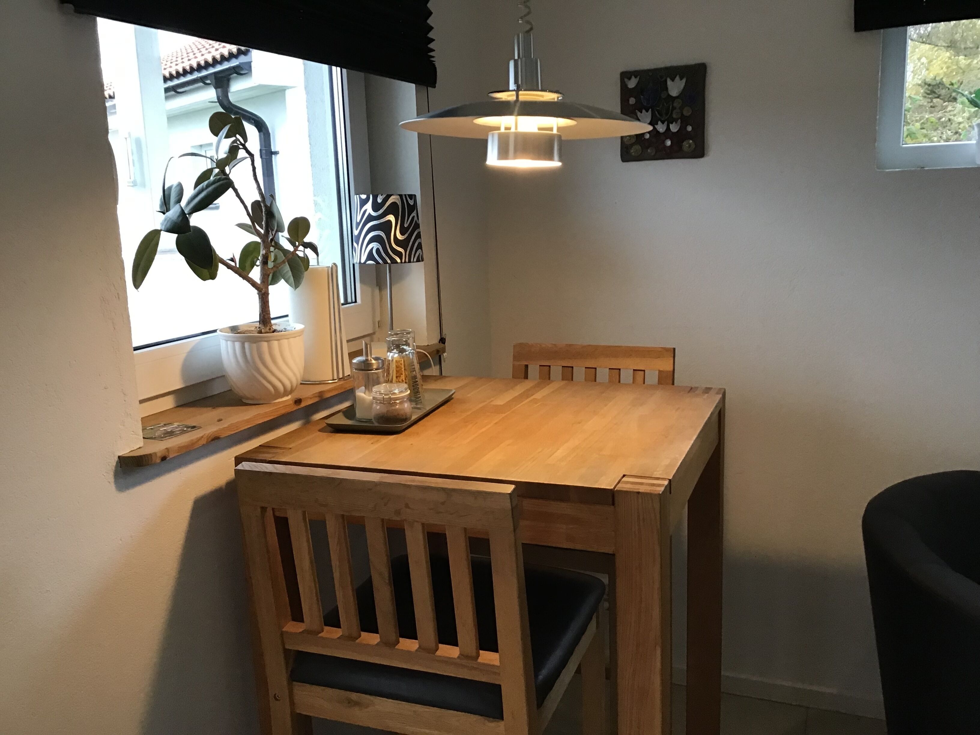 A wooden dining table with two chairs, a hanging light fixture, a potted plant, and a wall-mounted picture.