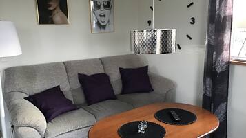 Una sala de estar con un sofá gris, dos alfombras negras, una mesa de centro de madera, un reloj de pared y una lámpara.